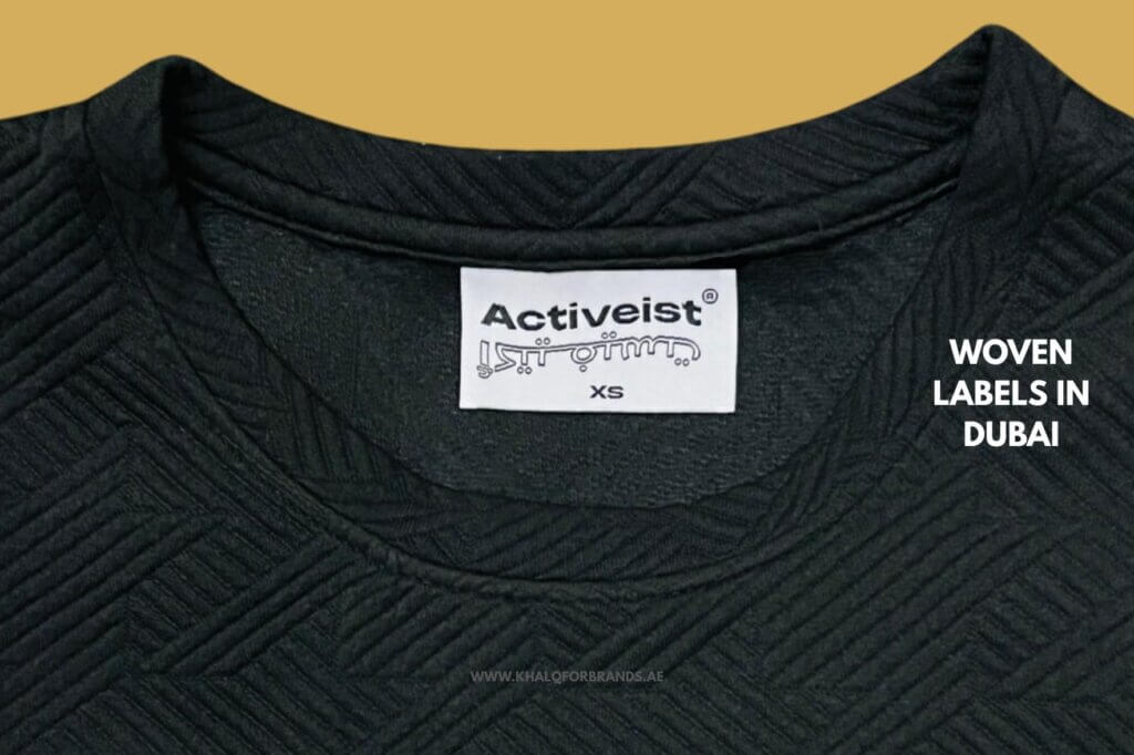 This image shows a black textured garment featuring a neatly stitched white woven label with the brand name Activeist. Clothing brands in Dubai use these custom woven labels to enhance product quality and branding.