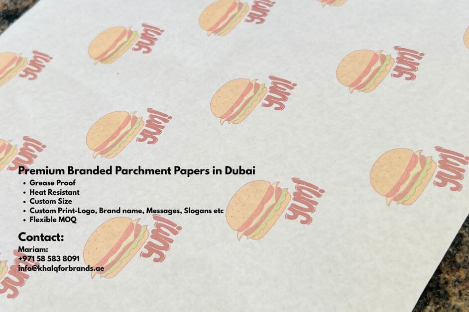 A close-up of a sheet of custom-printed parchment paper decorated with repeating burger illustrations and the word 'yum' in red. On the left side, black text highlights product details, reading 'Premium Branded Parchment Papers in Dubai' followed by bullet points: grease proof, heat resistant, custom size, custom print with logo, brand name, messages, slogans, and flexible MOQ. Below, contact details are provided: Mariam, phone number +971 58 583 8091, and email info@khalqforbrands.ae