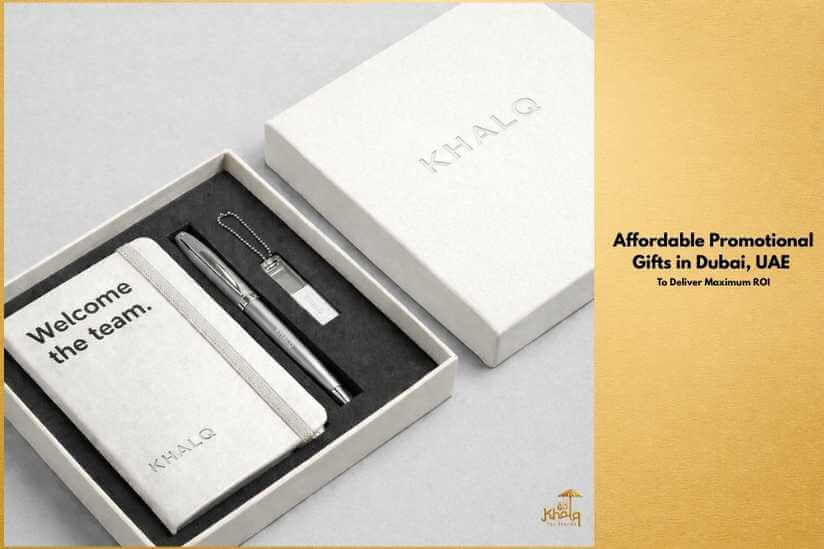 A premium corporate gift box displayed on a light background, containing a branded notebook, a metal pen, and a keychain neatly arranged inside a white box with the Khalq logo. The notebook reads “Welcome the team.” Text on the right side of the banner says “Affordable Promotional Gifts in Dubai, UAE – To Deliver Maximum ROI,” highlighting professional, budget-friendly corporate gifting.
